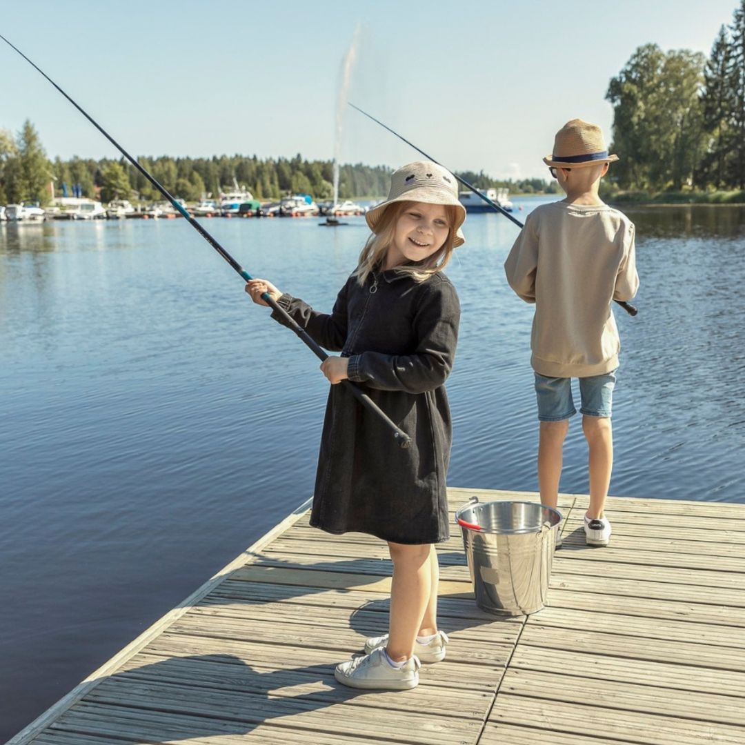 Kaksi lasta seisoo laiturilla järvessä, toinen on onkimassa, vieressä ämpäri ja aurinkoinen sää.