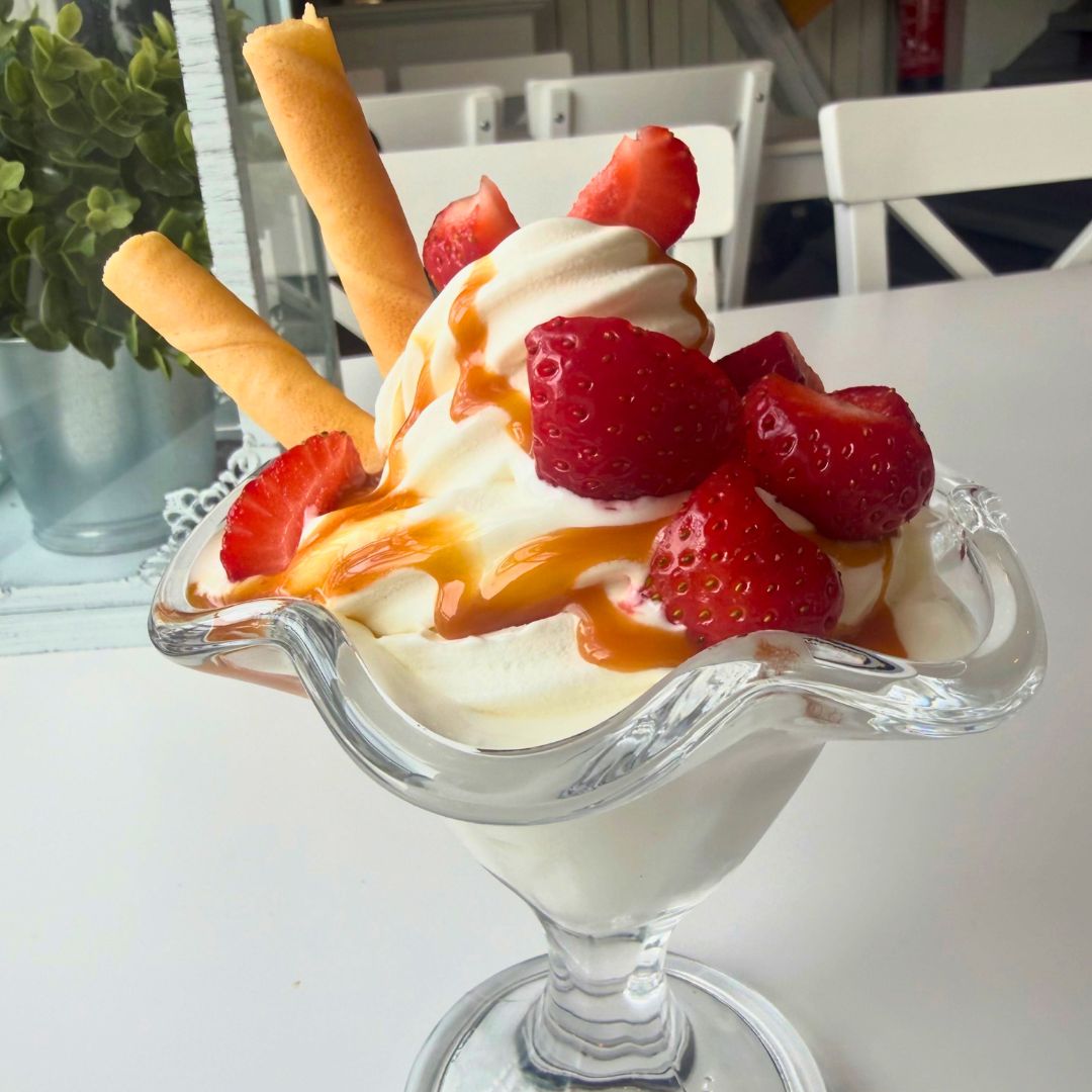 Eisbecher mit Vanille, Schokosauce, Streuseln, zwei Keksen, orangefarbener Blume und Löffeln auf Tisch.