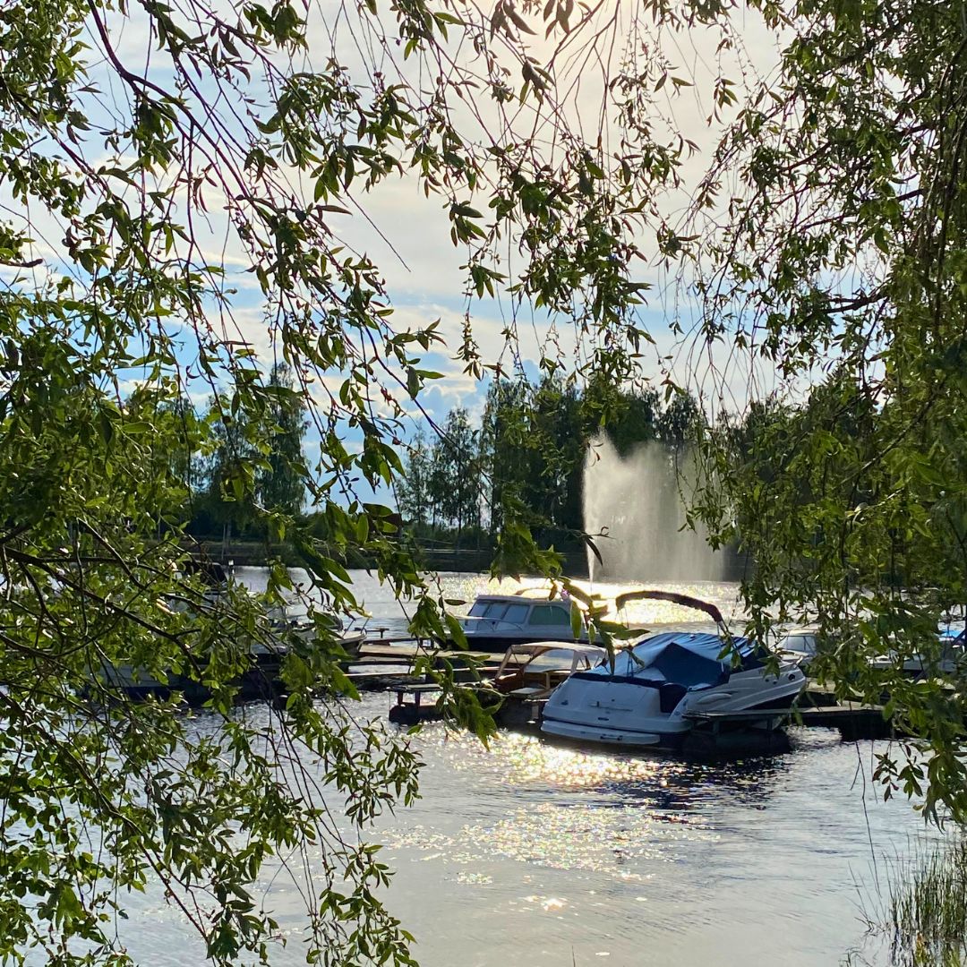 Järvimaisema, laitureita ja veneitä, suihkulähde suihkuttaa vettä, lehtien kehystämänä auringonpaisteessa.